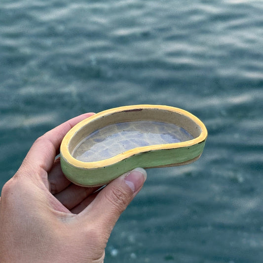 A hand holding a small, pool shaped ceramic dish or cup with a light green body and a yellow rim, set against a blurred background of rippling blue-green water.