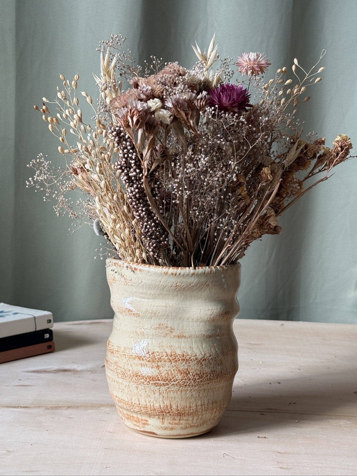 Decorative textured ceramic vase with dried flowers on a wooden surface against a green curtain background