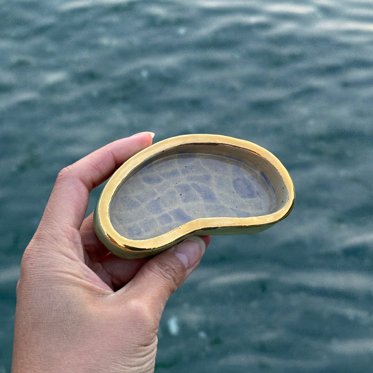 A hand holding a small, pool shaped gold-framed glass dish with a textured, patterned interior, set against a background of rippling blue-green water.
