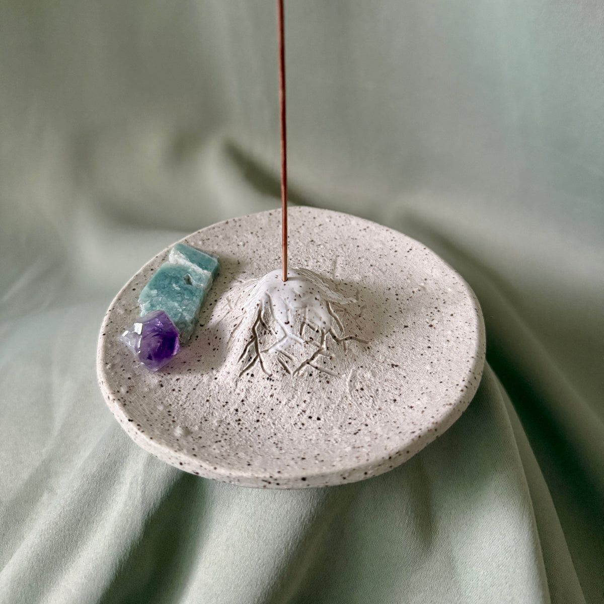 A round speckled ceramic incense holder with a mountain-shaped top and an incense stick burning from the peak, styled with two small crystals on a draped sage green fabric background.