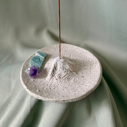 A round speckled ceramic incense holder with a mountain-shaped top and an incense stick burning from the peak, styled with two small crystals on a draped sage green fabric background.
