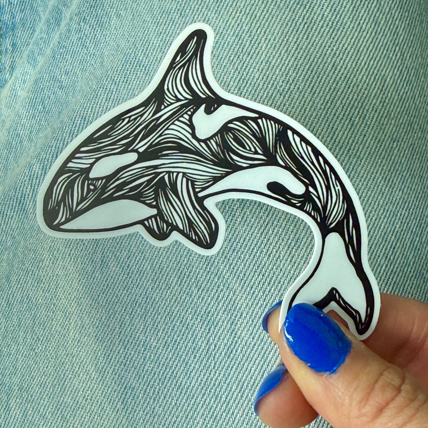 Hand holding a black and white stylized orca whale sticker on denim fabric