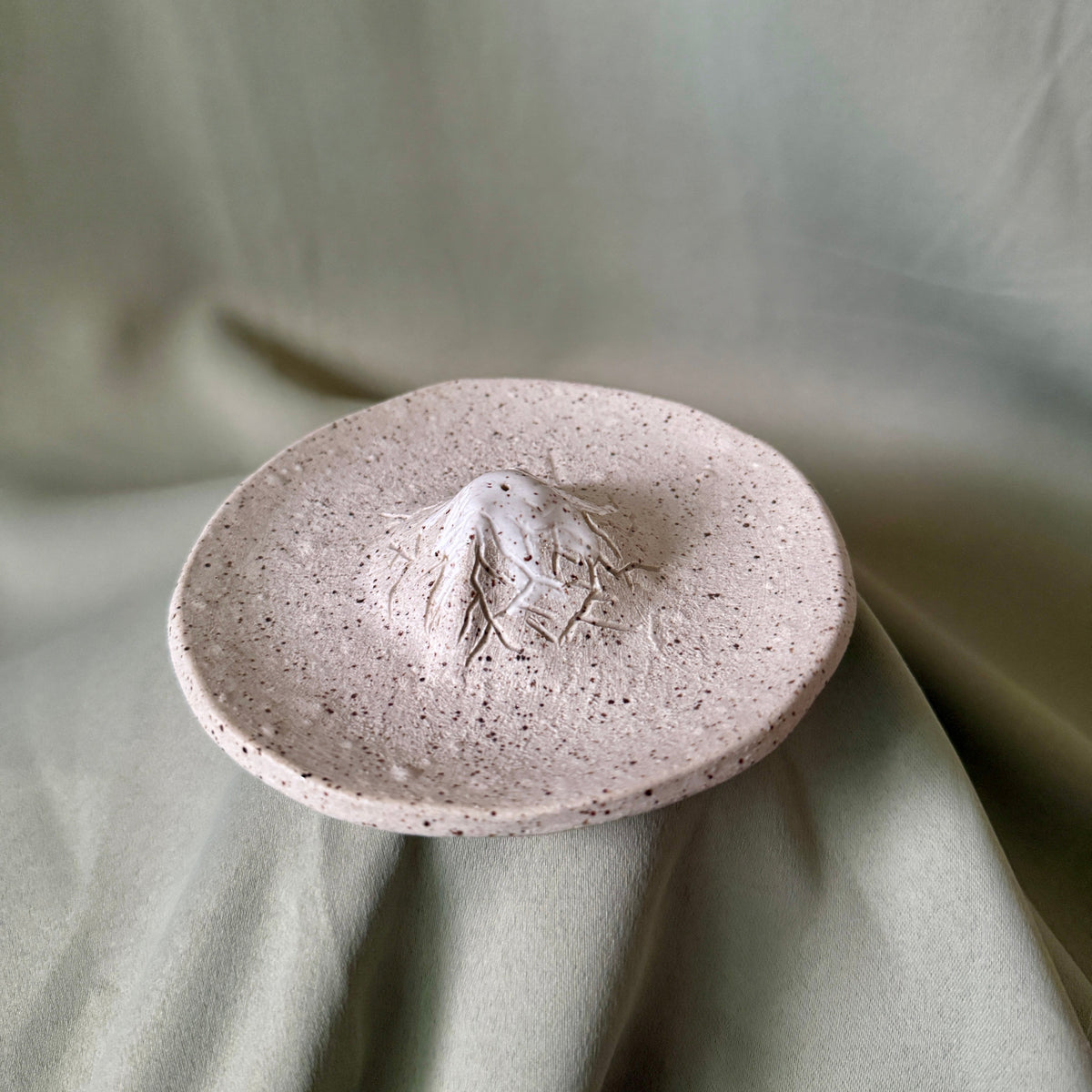 A small round ceramic dish with a textured speckled finish and a raised central mountain-like relief, shown on draped sage green fabric.