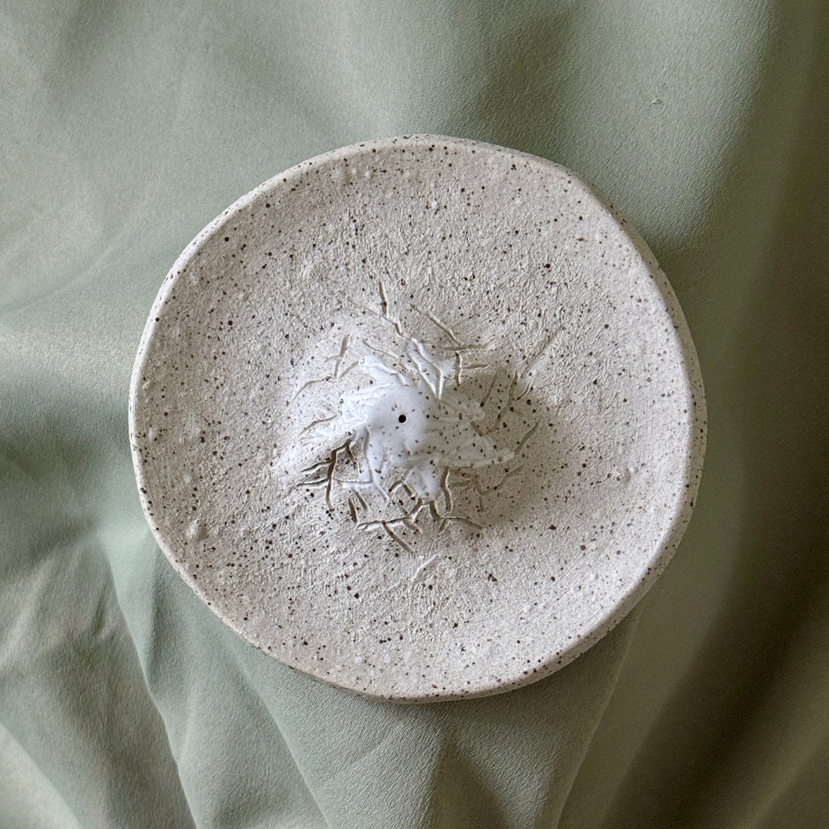 A round, handmade ceramic dish with a textured, speckled off-white surface and a small raised floral relief in the center, displayed on draped sage green fabric.