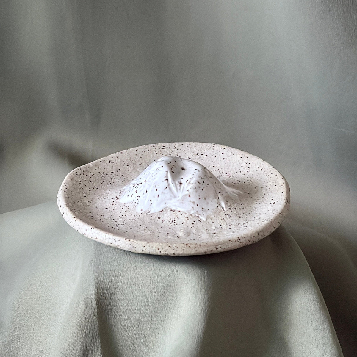 A small handmade ceramic dish with a textured, speckled off-white glaze, featuring a central raised peak design. The dish is displayed on draped sage green fabric against a matching fabric backdrop.