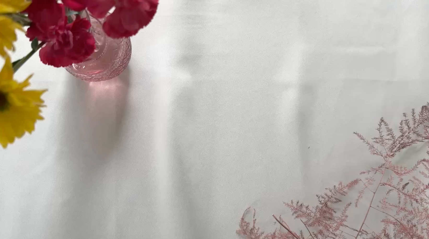 A white textured fabric backdrop with dried pink flowers in the bottom right corner and a small pink glass vase with yellow and red flowers in the top left, leaving open space in the center.