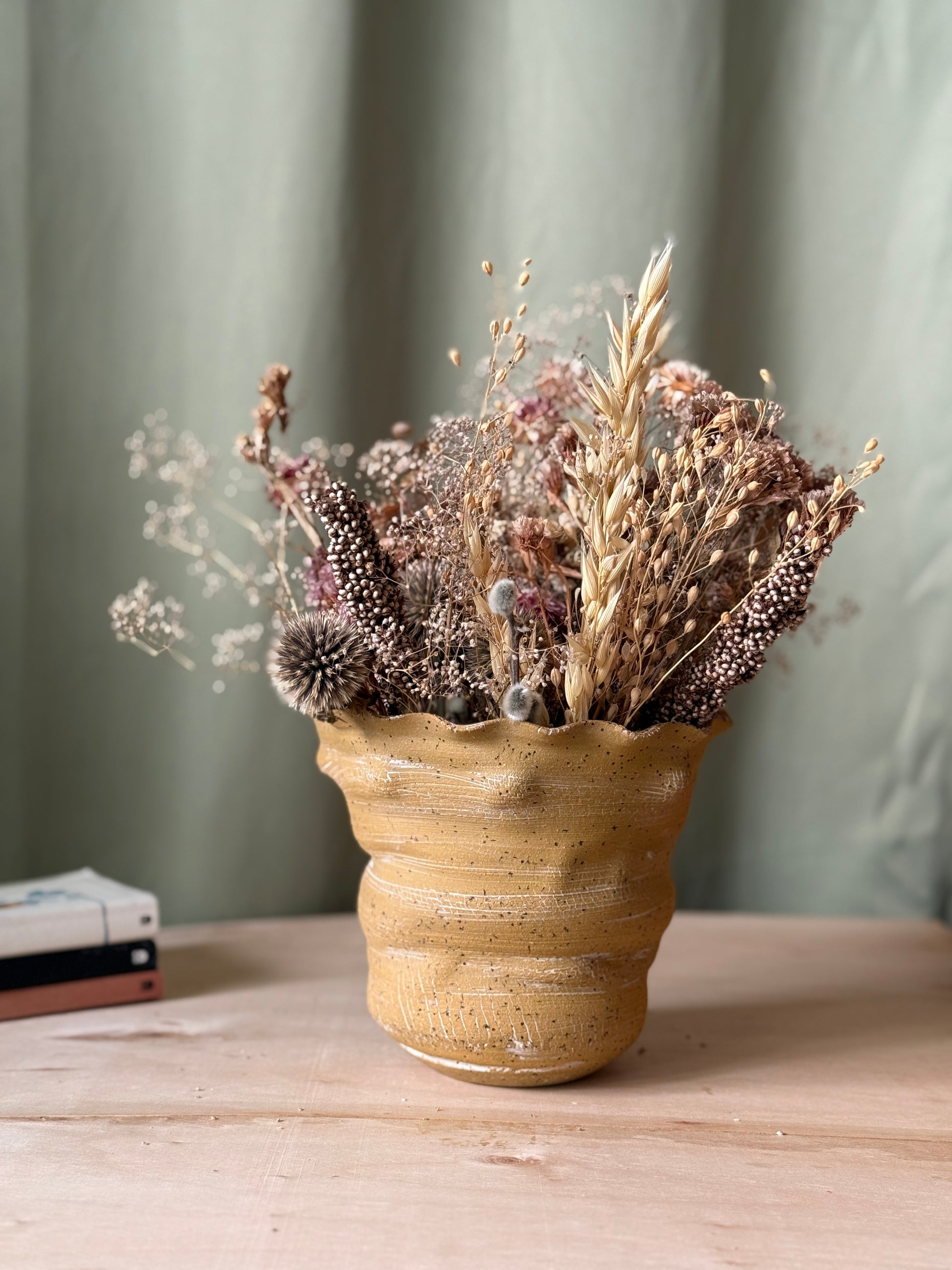 Ceramic texured vase with dried flowers on a wooden surface against a green curtain background