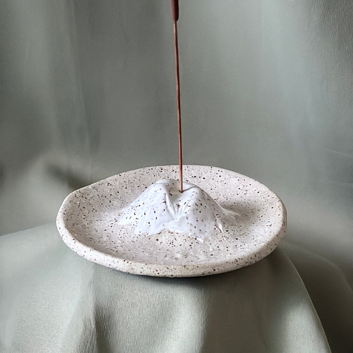 A small speckled ceramic mountain-shaped incense holder with a mountain peak and a vertical incense stick inserted, set against draped light gray fabric.