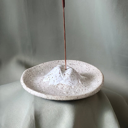 A small speckled ceramic mountain-shaped incense holder with a mountain peak and a vertical incense stick inserted, set against draped light gray fabric.