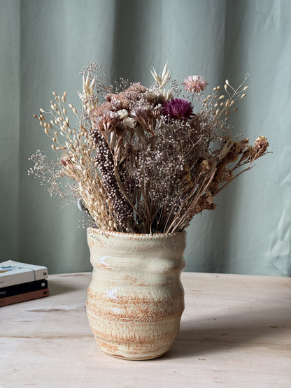 Decorative textured ceramic vase with dried flowers on a wooden surface against a green curtain background