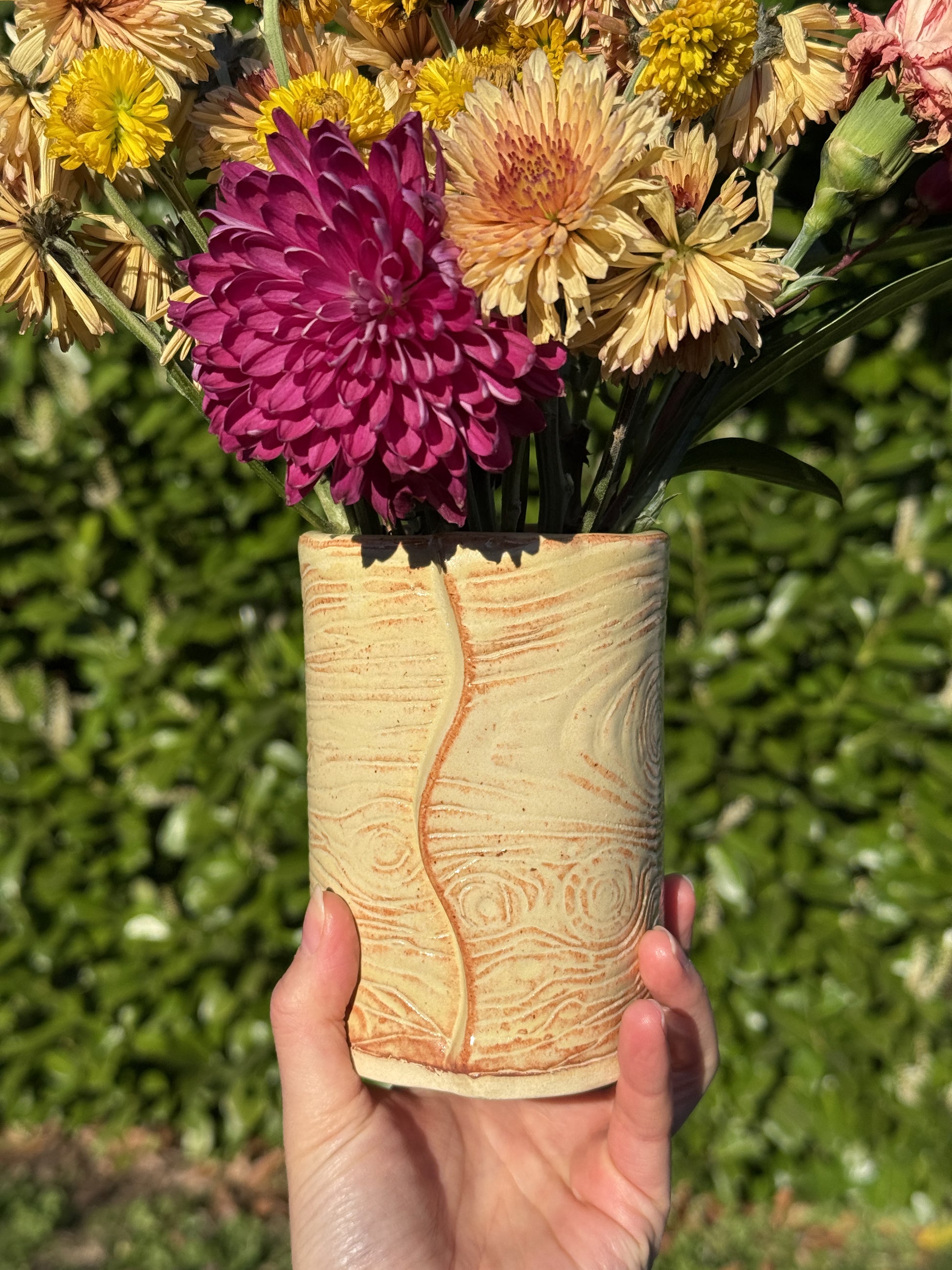 Hand holding a cylindrical wod grain ceramic vase with flowers in the foreground