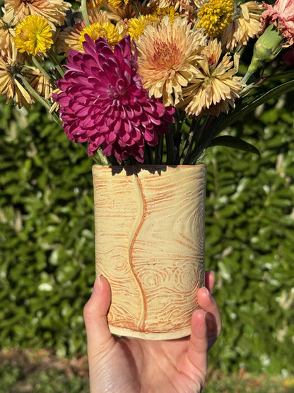 Hand holding a cylindrical wod grain ceramic vase with flowers in the foreground